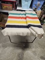 Blanket folded on metal stand showing colorful stripes in green, orange, yellow, and black on cream wool background.