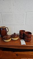 Full set showing two covered crocks, four brown bowls, and a pitcher arranged on a wooden surface against a white brick wall.