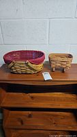 Overview of both baskets side by side on a wooden surface against a light wall. The left basket is oval with a red fabric liner, the right basket is a smaller cradle basket.