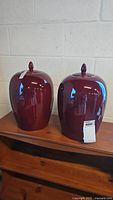 Two burgundy ceramic decorative jars with lids sitting on a wooden surface against a white wall.