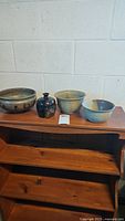 Four pottery pieces displayed on a wooden shelf including a small dark vase with stopper, two bowls, and a larger bowl with a mottled glaze.