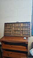 Wide shot of the primitive wooden printer's drawer resting on a wooden shelf, showing wear and multiple compartments.