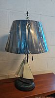 Metal sailboat table lamp with plastic-covered dark blue-gray lampshade and anchor pull chain.