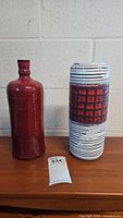 Front view of two vases; one red glossy vase and one white/black/red patterned Scheurich vase with a lot number card nearby