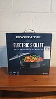 Front view of the Ovente electric skillet box showing the product name, 12-inch skillet with tempered glass lid, and food image on the skillet.