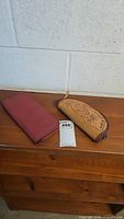 Two leather items on wooden surface against white wall: red leather folded organizer and tan clutch with floral stitching