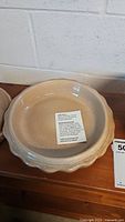 Photo of vintage beige pottery pie dish showing the scalloped edges and the flat inside base with a label inside. The piece is on a wooden surface against a concrete wall.