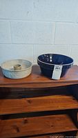 Two ceramic bowls: one white speckled bundt pan and one deep blue bowl side by side on wooden shelf against white brick wall.