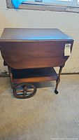 Front view of vintage wooden drop leaf tea cart, showing folded drop leaf and one large wooden wheel on rear side.