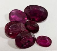 Photo showing six oval faceted ruby gemstones of various sizes, deep red color with visible inclusions on white background.
