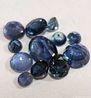 Close-up of twelve genuine blue sapphire gemstones in various faceted oval and round shapes, arranged on a white surface.