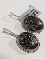 Close-up photo of two oval black druzy stones set in polished silver earrings with hook clasps, showing texture and sparkle of druzy.