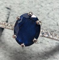 Close-up front angled view of the ring showing the oval blue sapphire and part of the silver band with clear accent stones.