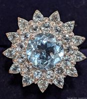Close up of silver ring top down showing round blue topaz center with surrounding clear stones in floral petal arrangement on black background.