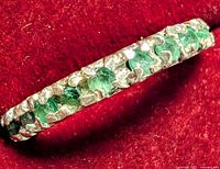 Close-up photo of silver full eternity ring with emerald stones on red velvet background, showing top side detail of stones and metal setting.