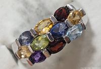 Close-up angle of silver ring front showing multi colored faceted gemstones in marquise and oval cuts placed in three rows.