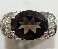 Close up front view of ring showing large dark brown faceted oval center gemstone and surrounding light green gemstones on silver band.