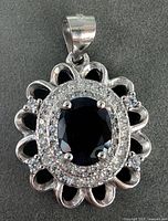 Silver pendant with a 2-carat oval sapphire surrounded by a halo of small diamonds, featuring a scalloped silver outer design and marked bail.