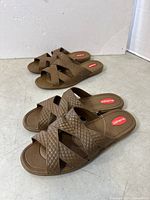 Two pairs of large brown Okabashi slippers with crossed straps seen together on a concrete floor.