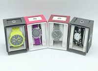 Four wristwatches in original packaging displayed upright: yellow watch, purple watch, silver-toned watch, and black watch with decorative strap.