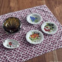 Top view of lot including amber glass ashtray and five ceramic dishes with fruit and floral designs arranged on pink geometric cloth