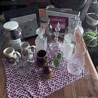 Photo of assorted glass decanters, pitchers, jars, ceramic mugs, and boxed drinking games on patterned cloth