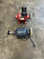 Top-down view of two bench grinders on a concrete floor. Red grinder with grinding wheel and protective cover; blue grinder with grinding wheel and wire brush attachment.