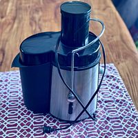 Front view of stainless steel Big Boss juicer with black plastic container and power cord laid on a patterned cloth.