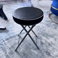 Side angle view of the black folding stool showing the X-shaped metal legs and round seat with white trim.