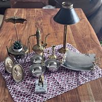 Full assortment of brass and metal household items displayed on patterned cloth on wooden table. Items include planter with umbrella, brass teapot, candle holder, table lamp, bowls, plaques, and tray.