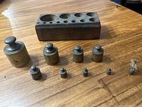 Photo of eight brass weights of varying sizes arranged in front of a rectangular wooden holder with compartments.