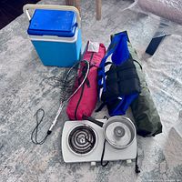 Photo of camping lot showing cooler, fishing net, red tent bag, life jacket, green mattress bag, and two-burner electric stove with pot.