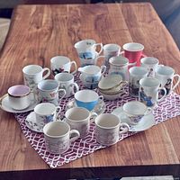 Overview of 22 assorted coffee and tea cups and some saucers on a wooden table with a purple patterned cloth