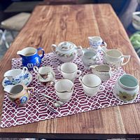 Overall lot of assorted vintage cream and sugar pieces laid out on a red and white geometric patterned cloth on wooden table