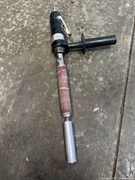 Custom air-powered sanding tool laid on the floor, showing full length with sanding drum and black pistol grip handle with throttle lever and side handle.
