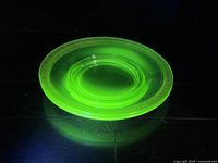 Single green Vaseline glass plate glowing vividly under UV light highlighting floral pattern and gold trim.