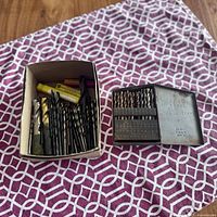 Box of mixed drill bits of various sizes and coatings alongside a metal case with smaller drill bits arranged inside.