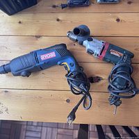 Both Ryobi drill and Makita hammer drill placed side by side on a wooden surface, corded and showing handles and part of the cords.