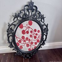 Front view of vintage black plastic circular frame with ornate floral and scroll design, containing glass and backing with red and white art inside.