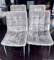 Four grey velvet dining chairs arranged in two rows on a dark wooden floor. Each chair has a tufted square pattern on the seat and backrest, with slim black metal legs.