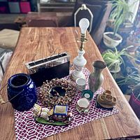 Multiple assorted decor items arranged on a patterned cloth on a wooden table including lamp base, wreath, blue lantern, carriage figurine