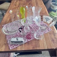Wide view of assorted glassware including various bowls, vases, and decorative glass pieces arranged on a patterned cloth on a wooden table.