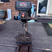 Front view of Jobmate drill press showing on/off switch and drill bit in chuck mounted on metal base with slots