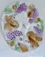Clear glass serving platter with raised embossed purple grapes, brown leaves, green accents. Scalloped edges with textured orchard design.