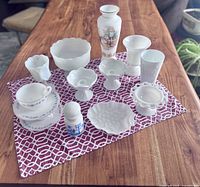 Full lot assortment displayed on wooden table showing all milk glass items including teacups, pedestal dishes, vase, candle holders, decorative plate, and jar.