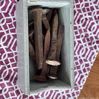 Top-down view into a small wooden box holding 10 rusty railway spikes with visible wear and rust.