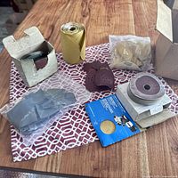 Wide view of assortment of sand paper sheets, sanding discs on box, sanding gloves, sanding pads, and small sanding accessories bag on table.