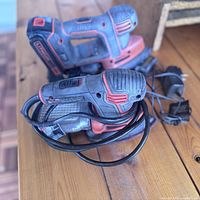 Photo showing the battery-operated sander and corded sander, both red and black, placed on a wooden surface with their cords visible.