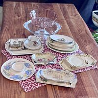 Full lot of assorted vintage ceramic platters, pink glass bowl, cup and saucer set on wooden table with red and white patterned cloth.
