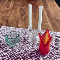 Full view of all four glass items displayed on a patterned cloth. Includes two white taper candles in a pair of clear glass holders, a green glass candle holder, and a red decorative glass piece.